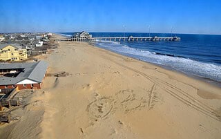 Beachfront view with sand, ocean waves, and a pier at Daniels' Homeport Coastal Furnishings.